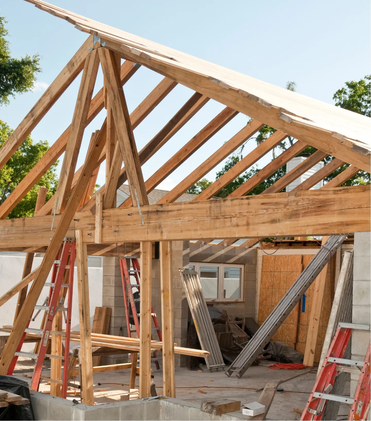 Wooden frame of a house under construction with ladders, beams, and tools visible on a sunny day.