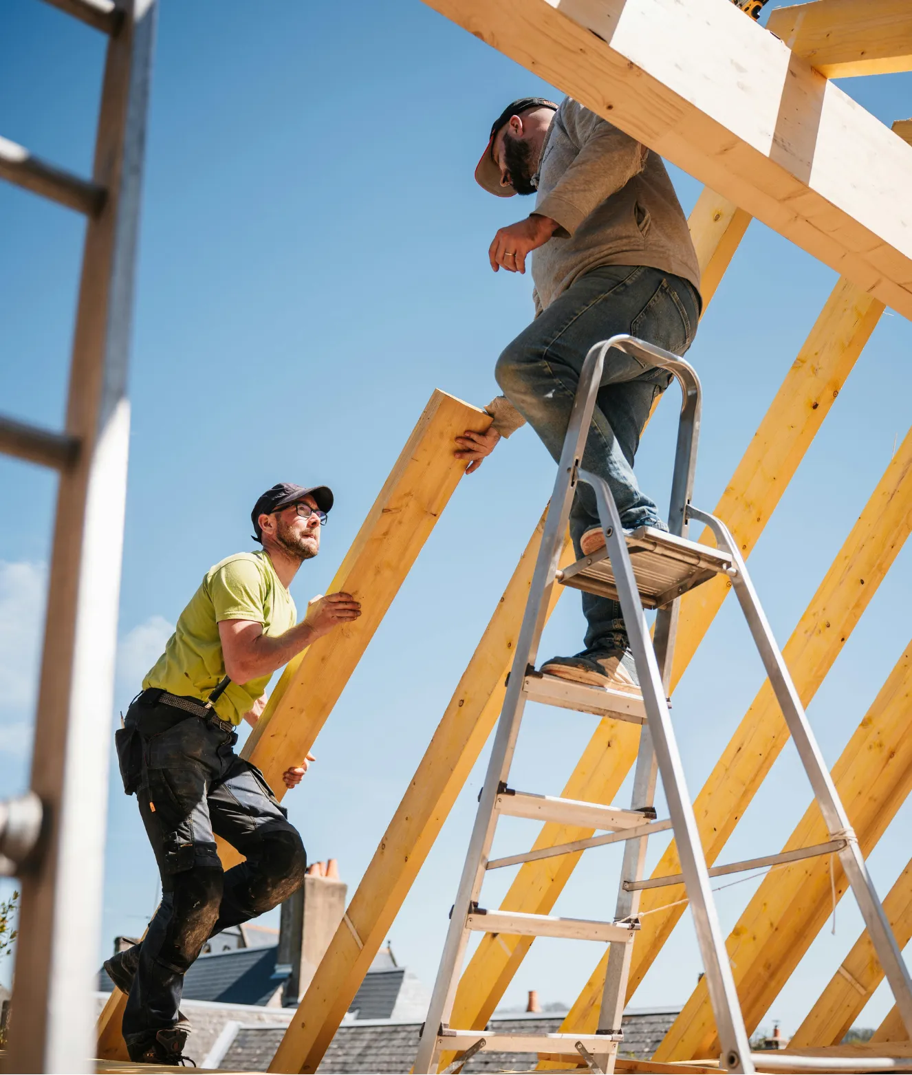 Two builders work together on a wooden roof frame; one stands on a ladder, the other passes a beam.