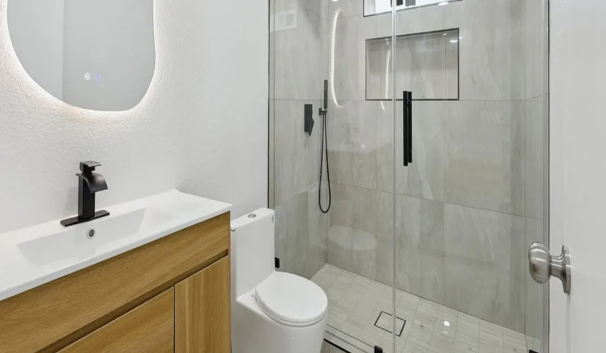 Modern bathroom with wood vanity, white sink and toilet, and glass shower with black fixtures.