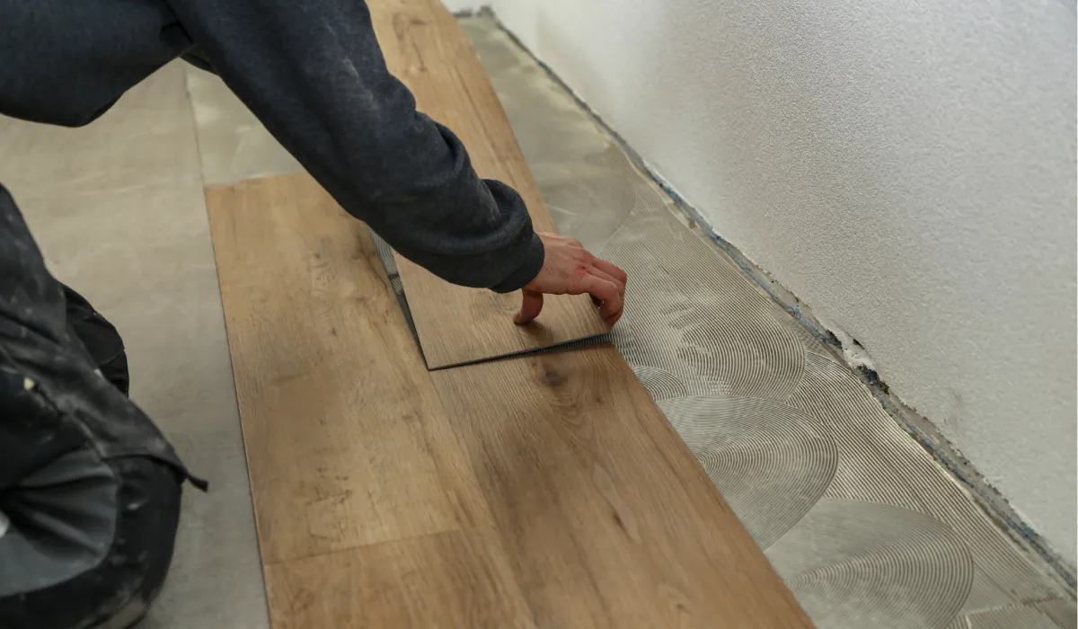 Person installing wooden flooring panels on a floor beside a white wall.