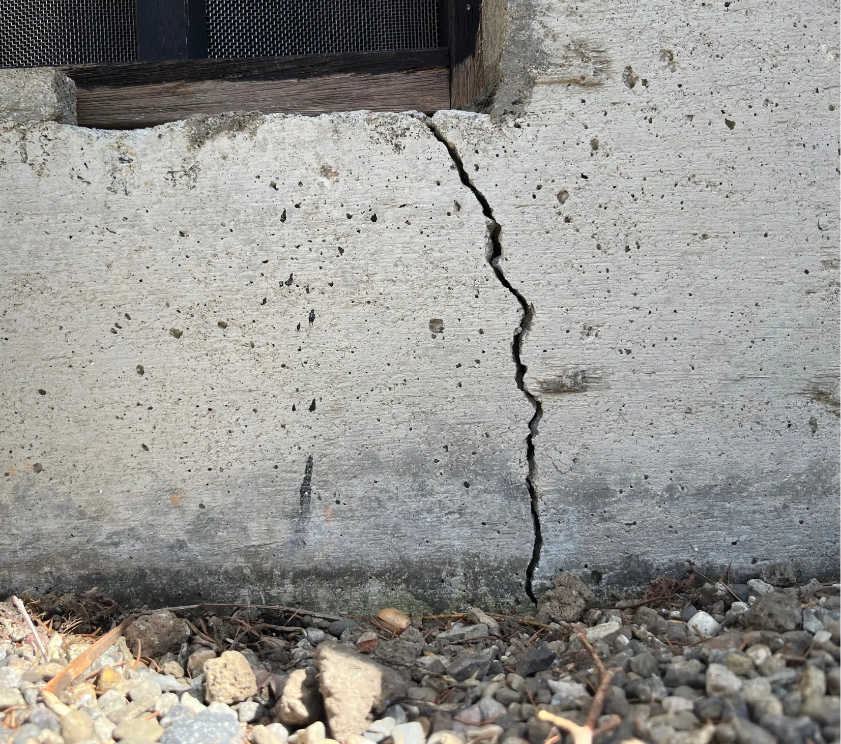A vertical crack runs up a concrete wall near the ground, with gravel and small rocks at the base.