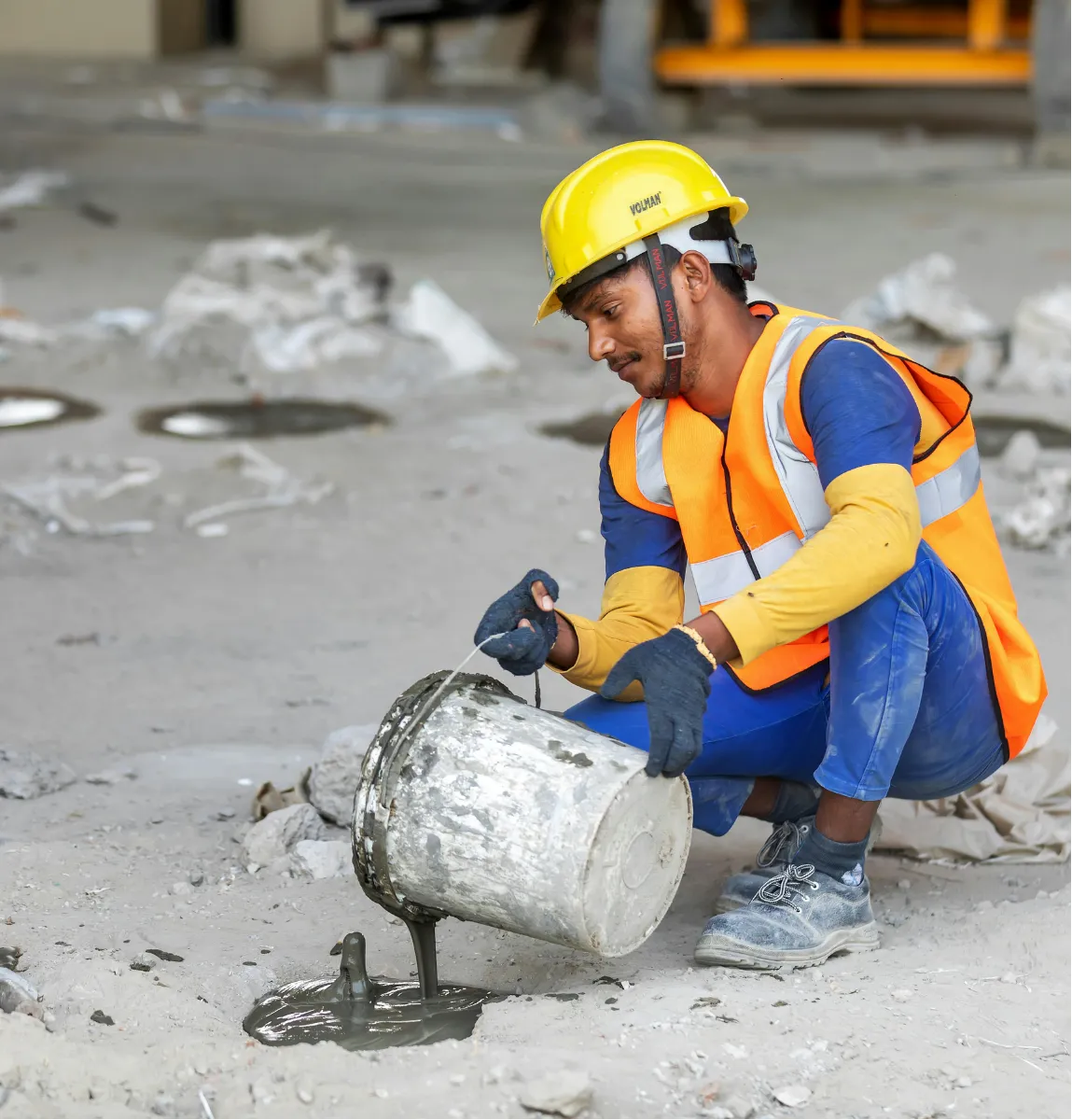 Construction worker in safety gear pours cement from a bucket onto the floor at a work site.