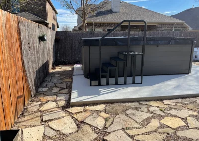 A backyard with a stone walkway, wooden fence, and above-ground pool with black stairs on a concrete pad.