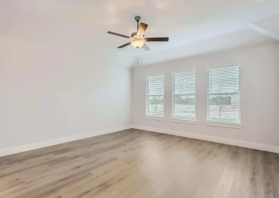 Empty room with light wood floors, three large windows with blinds, and a ceiling fan with lights.
