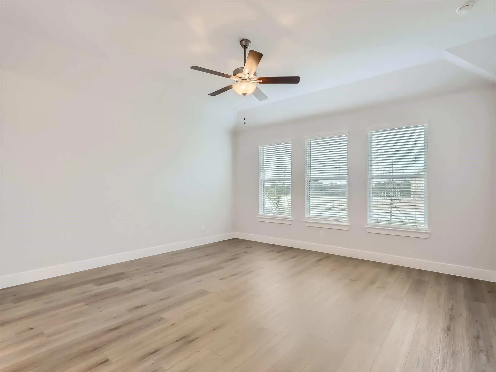 Empty room with light wood floors, three large windows with blinds, and a ceiling fan with lights.