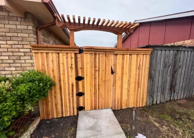 A wooden gate with a pergola top between two fences, next to a brick wall and bushes.