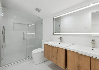 Modern bathroom with glass shower, double sinks with wood cabinets, and large illuminated mirror.