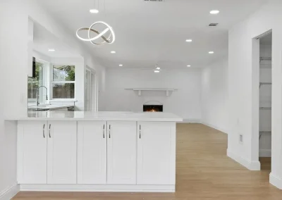 Modern white kitchen with island, open to a bright living area with fireplace and wood flooring.
