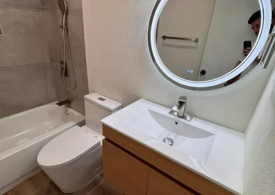 Modern bathroom with wood floor, bathtub-shower combo, wall-mounted sink, round lit mirror, and toilet.