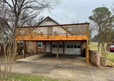 A house with a double garage and a large wooden deck built above the garage, surrounded by trees.