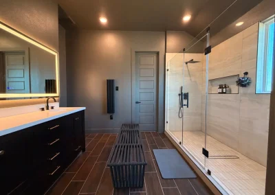 Modern bathroom with large glass shower, double sink vanity, bench, and dark wood-style flooring.