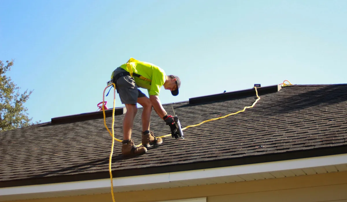 A worker in safety gear uses a nail gun while installing shingles on a sloped residential roof.