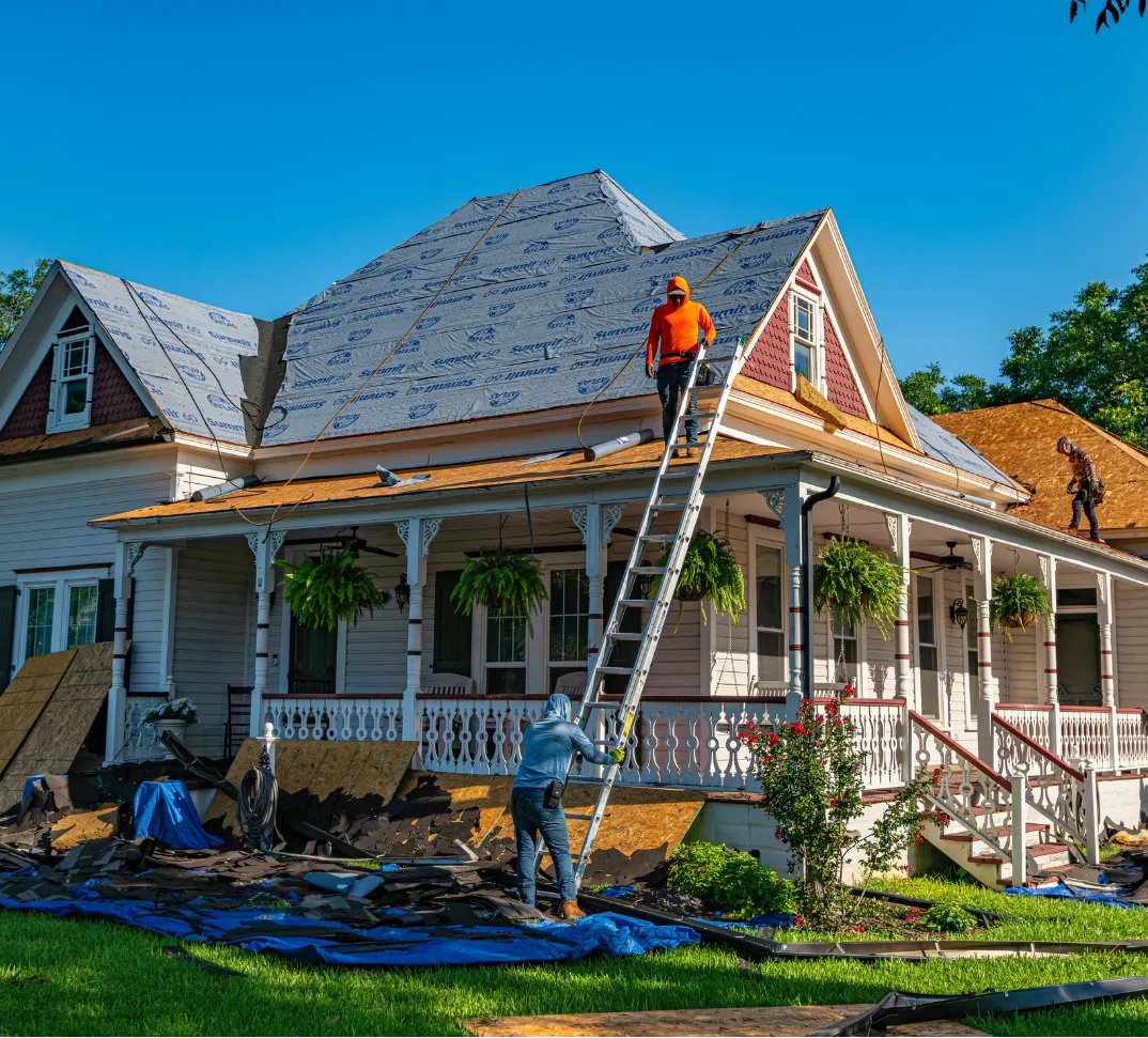 roofing-3 Two workers replace the roof of a house using ladders, with old shingles and debris on the ground.
